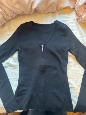 Svart ribbad långärmad topp - Svart långärmad topp med ribbad struktur och v-ringning med dragkedja framtill. Snygg och enkel modell som passar till mycket. Materialet känns mjukt och stretchigt. Endast testad en gång