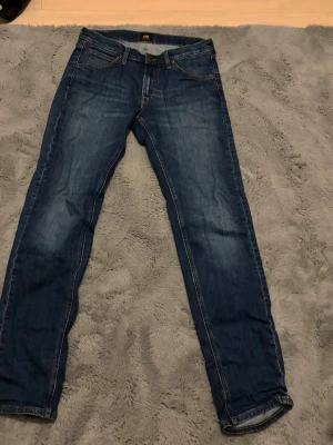 Blå jeans från Lee med raka ben - Säljer ett par klassiska blå jeans från Lee med raka ben och snygga slitningar. Jeansen har fem fickor, gylf med dragkedja. Ny pris ca 1200kr