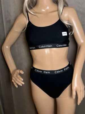 Svart Calvin Klein underklädesset M - Snyggt svart underklädesset från Calvin Klein med sportig topp och matchande trosor. Båda delarna har elastiska band med logotypen 'Calvin Klein' i vitt. Materialet känns mjukt och stretchigt, perfekt för en bekväm känsla hela dagen.