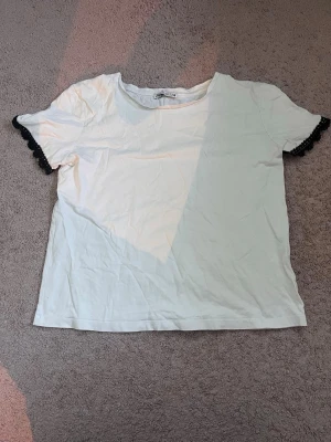 Vit t-shirt med spetsdetalj - Vit t-shirt i bomull med korta ärmar och svarta spetsdetaljer vid ärmsluten. Enkel och stilren design med rund halsringning. Perfekt för dig som gillar en basic look med en twist.