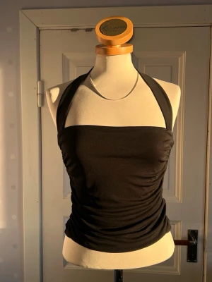 Svart halterneck topp S - Snygg svart halterneck topp med åtsittande passform och breda axelband. Toppen är ärmlös och har en croppad look som passar perfekt till både jeans och kjol. Tillverkad i mjukt, stretchigt material för en bekväm känsla.