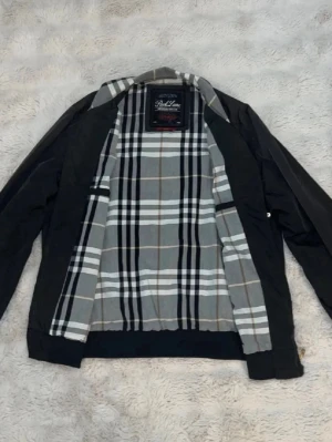 Svart bomberjacka med rutigt foder - Snygg svart bomberjacka från Red Line med klassiskt rutigt foder i grått, vitt och svart. Jackan har dragkedja framtill, ribbade muddar vid ärmslut och midja samt två fickor med dragkedja. Perfekt för dig som vill ha en stilren och tidlös look.