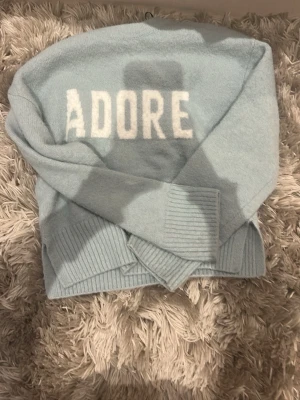 Ljusblå stickad tröja med text - Mysig ljusblå stickad tröja med vit texten 'ADORE' framtill. Tröjan har långa ärmar och breda ribbade muddar vid ärmslut och nederkant. Perfekt för dig som gillar en enkel men trendig look.