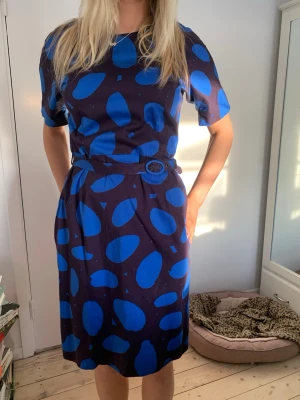 Blåmönstrad kort klänning med bälte från Marimekko - Midiklänning i mörklila med stora blåklintsblå prickar och matchande bälte i midjan. Klänningen har rund halsringning och korta ärmar och en stram passform som gör den mycket användbar. Perfekt för dig som vill sticka ut med både färg och mönster i klassisk Marimekko-stil. Två pyttesmå fläckar direkt vid urringningen, se bild. Modellen har storlek 36/38 och är 1,75 lång.