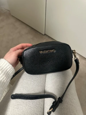 Svart crossbody från Valentino - Snygg svart handväska från Valentino i crossbody-modell. Väskan är gjord i strukturerat läder med guldfärgade detaljer och dragkedja. Den har justerbar axelrem och ett rymligt huvudfack med innerficka. Perfekt för att bära det viktigaste med stil.