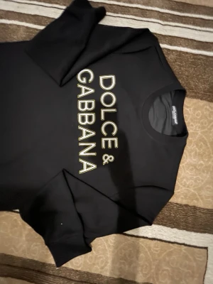 Dolce Gabbana Hoodie - Snygg svart t-shirt från Dolce & Gabbana med stort vitt och gult logotryck på bröstet. Klassisk rund halsringning och korta ärmar. Tillverkad i mjukt material som känns skönt mot huden. Perfekt för dig som gillar stilrena designer med lyxig känsla.
