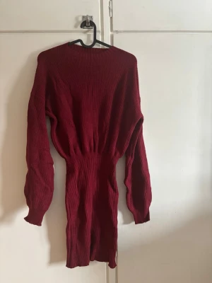 Vinröd ribbad stickad klänning - Mysig vinröd stickad klänning med ribbad struktur och lång ärm. Klänningen har en markerad midja och är i kortare modell, perfekt för lager på lager. Passformen är normal och materialet känns mjukt och stretchigt.