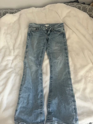 Ljusblå lowwaist bootcut jeans - Säljer ett par ljusblå lowwaist bootcut jeans med låg midja. Jeansen har en avslappnad passform över låren och utsvängda ben. Perfekta för dig som gillar en retro vibe och vill ha ett par bekväma jeans till din garderob. Storlek 158 från gina Young (jag har innerbensläng 74cm för referens) säljs då de är försmå i midjan(därför ser dragkedjan sne ut)