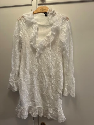 Vit spetsklänning med volang/ studentklänning  - Vit klänning i spets med v-ringning och volangdetaljer längs halsringningen och nederkanten, långärmad. Helt ny med prislapp! 