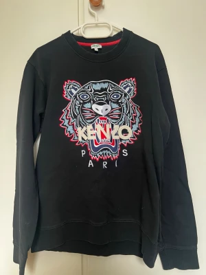 Svart sweatshirt med tiger från Kenzo - Säljer en svart sweatshirt från Kenzo med det ikoniska broderade tigerhuvudet och texten 'KENZO PARIS' på bröstet. Tröjan har rund halsringning, ribbade muddar och är tillverkad i mjukt bomullsmaterial. Perfekt för dig som vill sticka ut med en statement-piece.
