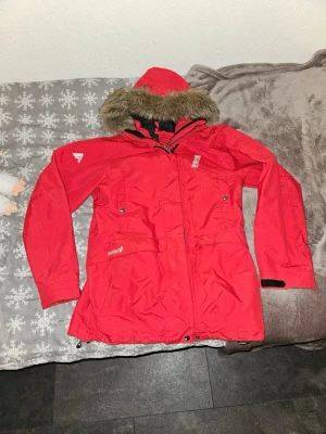Röd parkas från 8848 Altitude - Snygg röd parkas från 8848 Altitude med avtagbar huva och fuskpäls. Jackan har dragkedja, tryckknappar, flera fickor och justerbara ärmslut. Perfekt för kalla dagar och riktigt skön att bära. Märkeslogga på bröstet och ärmen.