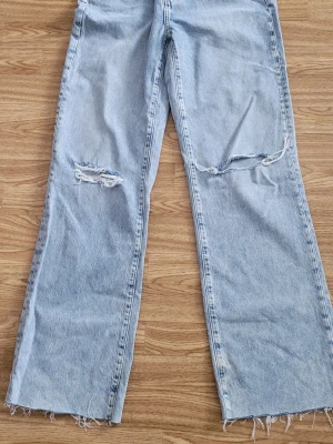 Ljusblå slitna raka jeans - Ljusblå jeans med raka ben och slitningar över knäna. Jeansen har råa, fransiga benslut och knäppning med silverfärgade knappar. Klassisk femficksmodell i denim med gul söm. Perfekta för en avslappnad och trendig look.