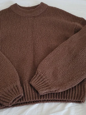 Brun stickad tröja med rund hals - Mysig brun stickad tröja med rund halsringning och breda ribbade muddar vid ärmslut och nederkant. Tröjan har en avslappnad passform och är tillverkad i ett mjukt, grovt garn som ger en skön känsla. Perfekt för kyliga dagar.