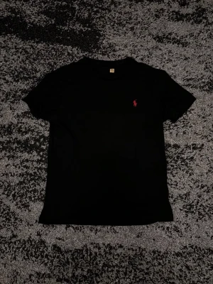 Svart t-shirt från Ralph Lauren - Klassisk svart t-shirt från Ralph Lauren med korta ärmar och den ikoniska röda logotypen broderad på bröstet. Enkel och stilren design i mjuk bomull som passar perfekt till all slags byxor.