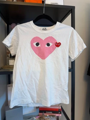 Vit t-shirt med hjärtmotiv från PLAY - Vit t-shirt från PLAY med ett stort rosa hjärta med ögon tryckt på bröstet och ett litet rött hjärta med ögon broderat bredvid. Klassisk rund hals och korta ärmar. En liten defekt.. vet ej om de går bort. ingen garanti. se bild 3💞💞💞