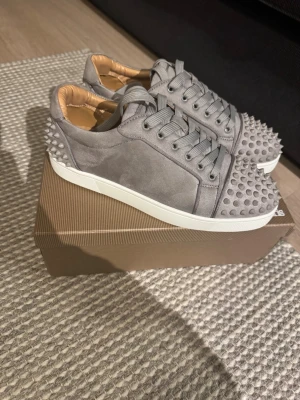 Gråa Christian Louboutin sneakers - Säljer ett par gråa sneakers från Christian Louboutin i mocka med ikoniska nitar över tå och häl. Skorna har vita sulor och den klassiska röda undersidan. Insidan är beige och de har matchande gråa skosnören. Perfekta för dig som vill sticka ut med exklusiv design.