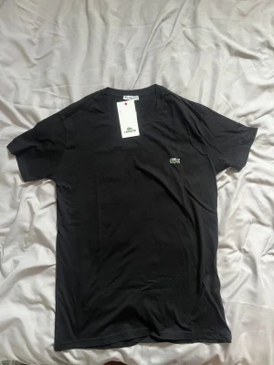 Svart t-shirt från Lacoste - Klassisk svart t-shirt från Lacoste med rund halsringning och det ikoniska krokodilmärket broderat på bröstet. T-shirten har korta ärmar och är tillverkad i mjuk bomull för en skön känsla. Perfekt basplagg för en clean och stilren look.