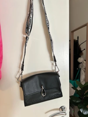 Svart axelväska Tommy Jeans - Snygg svart axelväska från Tommy Jeans med präglad logga framtill och coolt metallspänne. Väskan har en bred, justerbar axelrem med tydlig logotyp i svartvitt. Tillverkad i slätt konstläder och har en boxig, modern form.