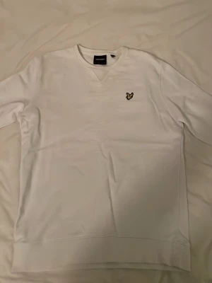 Vit sweatshirt från Lyle & Scott - Klassisk vit sweatshirt från Lyle & Scott med rund halsringning och den ikoniska gula örnloggan broderad på bröstet. Tröjan har en avslappnad passform och är tillverkad i mjukt bomullsmaterial. Perfekt för en clean och stilren look.