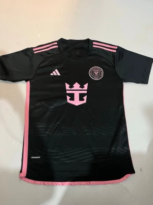 Svart Inter Miami matchtröja Adidas - Snygg svart matchtröja från Inter Miami med rosa detaljer och klubbmärke på bröstet. Tröjan har korta ärmar, rosa ränder på axlarna och rosa sidopaneler. Tillverkad i lätt och ventilerande material med Aeroready-teknologi.