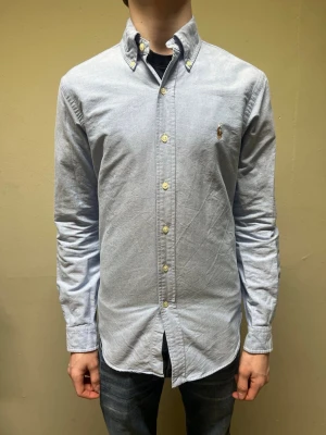 Ljusblå skjorta från Ralph Lauren - Klassisk ljusblå skjorta från Ralph Lauren med button-down krage och broderad logga på bröstet. Skjortan har lång ärm, normal passform och är tillverkad i mjuk bomull. Perfekt till jeans eller chinos för en clean look. Det är bara att höra av dig vid minsta intresse!