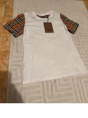 Vit Burberry t-shirt med rutiga ärmar - Säljer en vit t-shirt från Burberry med klassiska beige, röda och svarta rutiga korta ärmar. T-shirten har rund halsringning och är i mjuk bomull. Perfekt för dig som vill ha en ikonisk och stilren look med en twist.