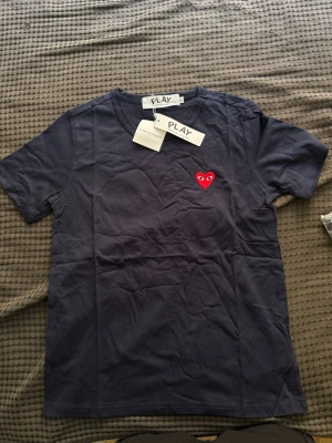 Mörkblå t-shirt Comme des Garçons Play - Snygg mörkblå t-shirt från Comme des Garçons Play med det ikoniska röda hjärtat broderat på bröstet. Klassisk v-ringad modell i mjuk bomull och korta ärmar. Perfekt för dig som gillar stilrena plagg med en twist.