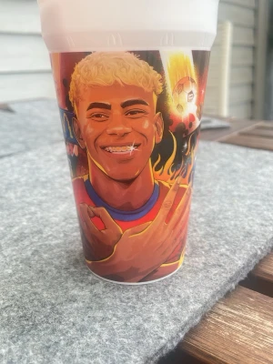 Plastmugg med Lamine Yamal-motiv - Cool plastmugg med färgstark illustration av fotbollsspelaren Lamine Yamal. Muggen har eldiga detaljer, tecknad fotboll och spelarens namn i grafisk stil. Perfekt för dig som gillar fotboll och vill ha något unikt till drycken.