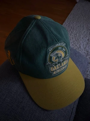 Grön och gul Oakland Athletics keps - Snygg keps från New Era i grönt och gult med broderad Oakland Athletics-logga framtill. Klassisk böjd skärm och justerbar passform. Tillverkad i bomullstyg och har detaljer som gul knapp på toppen och broderad New Era-logga på sidan.
