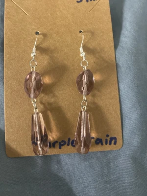 Lila droppformade örhängen - Snygga örhängen med två lila, facetterade pärlor i varje örhänge. Den övre pärlan är oval och den nedre är droppformad. Krokar i silverfärgad metall och transparenta pluppar baktill. Perfekta för dig som gillar unika accessoarer.