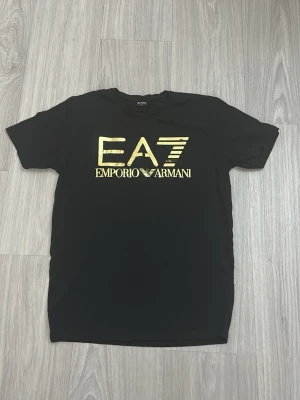 Svart EA7 Emporio Armani t-shirt - Snygg svart t-shirt från EA7 Emporio Armani med stort guldfärgat logotryck på bröstet. Klassisk passform med rund hals och korta ärmar. Perfekt för dig som vill ha en stilren och trendig look med exklusiv känsla.
