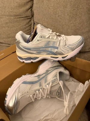 Vita och silver Asics sneakers - Snygga sneakers från Asics i vitt och silver med mesh- och syntetmaterial. Skorna har klassisk låg profil, vita snören och detaljer i ljusblått. Perfekta för dig som gillar sportig och clean stil.