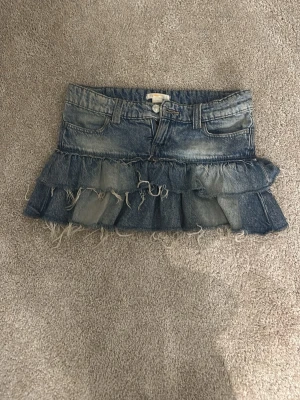 Jeanskjol med volanger från Gina Tricot - Säljer en blå jeanskjol från Gina Tricot med volangdetaljer och råa kanter. Kjolen har klassisk femficksdesign och knäppning fram. Perfekt för dig som gillar en cool och avslappnad stil.