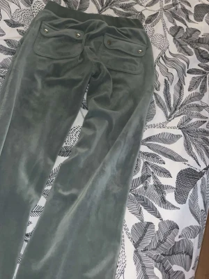 Grågröna velourbyxor med fickor - Mjukisbyxor i grågrönt velourtyg med bred resår i midjan och två stora fickor bak med knappdetaljer. Byxorna har raka ben och en avslappnad passform, perfekta för chill dagar. Snygg retrostil som passar till hoodie eller crop top.