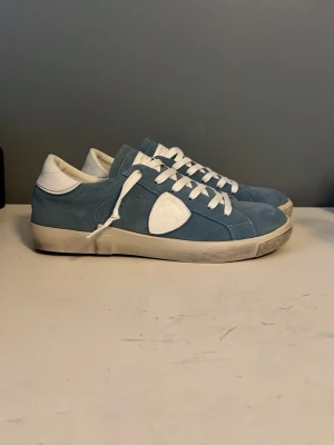 Blå Philippe Model sneakers i mocka - Snygga blå sneakers från Philippe Model i mjuk mocka med vita detaljer och vita skosnören. Skorna har en låg silhuett, vit sula och den klassiska sköldloggan på sidan. Perfekta för dig som gillar stilrena och trendiga sneakers. Helt oanvända, storlek 44, var lite små för mig så passar nog perfekt för 43! pris går att diskuteras. Låda och dustbag medföljer 