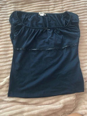 Mörkblå topp med spets och rosett - Söt mörkblå topp med rund halsringning, liten rosett framtill och spetsdetalj med band under bysten. Ärmlös modell i mjukt material, perfekt för en trendig och enkel look.