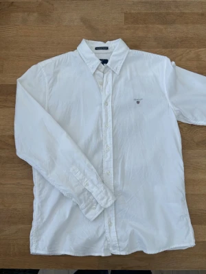 Vit skjorta från Gant med knappar - Klassisk vit skjorta från Gant med knappar framtill och broderad logga på bröstet. Skjortan har lång ärm, normal passform och är tillverkad i bomull. Perfekt till en clean och stilren look.
