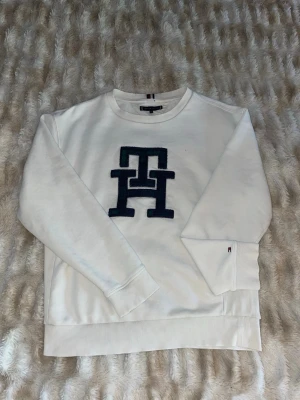 Tommy Hilfiger Sweatshirt - Vit sweatshirt från Tommy Hilfiger med stort blått TH-logotyp framtill. Tröjan har rund halsringning, ribbade muddar och diskret flaggdetalj på ärmen. Perfekt för en clean och stilren look. Limited edition
