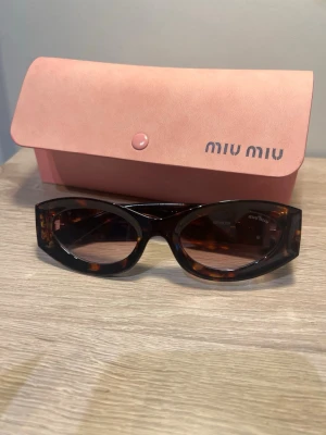 Bruna ovala solglasögon från Miu Miu - Snygga solglasögon från Miu Miu med oval form och brunmelerat mönster. Bågarna har en chunky design och är dekorerade med stora guldfärgade Miu Miu-loggor på sidorna. Kommer med ett rosa fodral med logga. Perfekt accessoar för en trendig look.