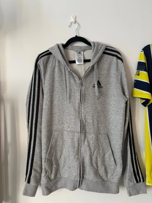 Grå zip hoodie från Adidas - Snygg grå hoodie från Adidas med dragkedja, huva och klassiska svarta ränder längs ärmarna. Adidas-logga på bröstet och två stora fickor framtill. Tillverkad i mjukt bomullsmaterial, perfekt för en avslappnad streetwear-look.