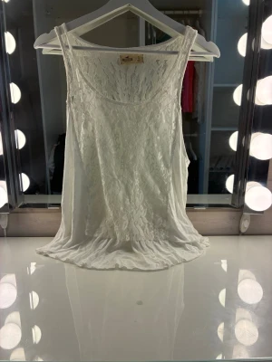 Vit spetslinnetopp från Hollister - Supersöt vit linnetopp från Hollister med blommig spets och rund halsringning. Toppen är ärmlös och har en flowy, lätt transparent look som passar perfekt till sommarens outfits.