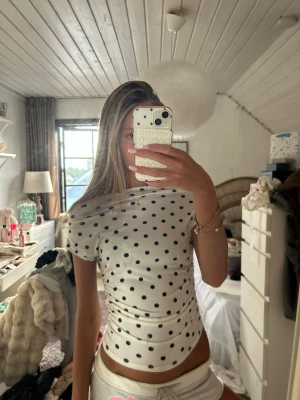 Vit prickig croppad topp - Säljer en vit croppad topp med svarta prickar. Toppen har korta ärmar och är figurnära med snygg rynkning i sidorna. Perfekt för dig som gillar trendiga och lekfulla plagg. Materialet känns mjukt och stretchigt.