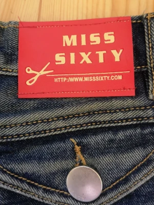 Miss Sixty blå jeans stl 26 - stl 26 midje 68cm  innerbenslängd 82 cm. längd 107cm. Material : 97% bomull  3% elestan  Använts ett fåtal gånger, i finfin skick, Jeans med fem fickor och hällor för bälte. Jeansen har en knappgylf och raka ben. Det finns detaljer som nitar och sömmar. Djur och rökfritt hem. Perfekt för dig som gillar en tidlös och trendig look.