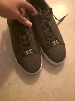 Bruna Michael Kors sneakers - Snygga bruna sneakers från Michael Kors med klassiskt MK-mönster och vita sulor. Skorna har bruna snören och metall-detaljer med MK-logga framtill. Tillverkade i ett tåligt material med låg profil och stilren design.