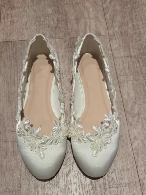 Satinskor i ballerinamodell med spetsdetaljer st 36 - Satinskor i Ballerinamodell med vacker spets och broderade blommor samt små pärlor längs kanten. Färg Ivory . Skorna har rund tå och är tillverkade i lyxig satin. Perfekta för dig som vill ha något extra till outfiten.