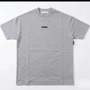 Grå Essentials T-shirt Fear of God - Snygg grå T-shirt från Fear of God Essentials med diskret logga på bröstet och stor präglad text på ryggen. Klassisk passform med rund hals och korta ärmar. Perfekt för en clean och stilren look.