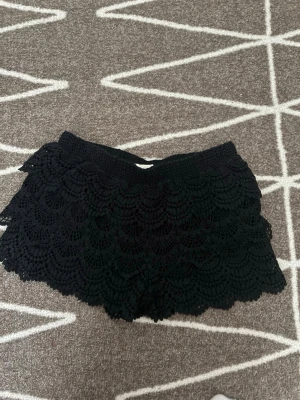 Svarta spets-shorts från Forever 21 - Svarta shorts från Forever 21 med snygga lager av spets och elastisk midja. Perfekta för sommaren med sin lätta och luftiga känsla. Ger en cool och trendig look med sin unika struktur och detaljerade spetsmönster. Storlek XS.🤍