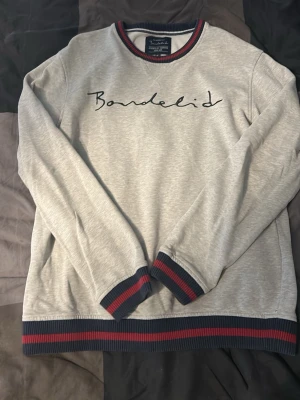 Grå långärmad topp Bondelid - Snygg grå långärmad topp från Bondelid med broderad logga framtill. Rund halsringning och muddar i marinblått och rött ger en sportig touch. Tillverkad i mjuk bomullsmix, perfekt för en avslappnad stil.
