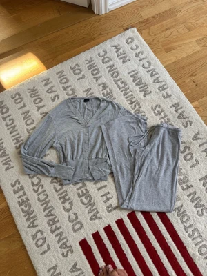 Grått loungewear-set från Gina Tricot - Mysigt grått loungewear-set från Gina Tricot med långärmad topp och matchande byxor. Toppen har v-ringning, knappar framtill och bred mudd i midjan för en skön passform. Perfekt för chill dagar hemma.