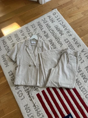 Randig linne matchande sett från Selected Femme - Säljer en beige och vit randig matchande set från Selected Femme. Modellen har korta ärmar, knytband i midjan och två knappar framtill. Tillverkad i ett lätt och luftigt material, perfekt för en avslappnad och trendig look. Toppen är i storlek 40 och byxorna är i storlek 38. Men tycker att den passar någon som är storlek M 
